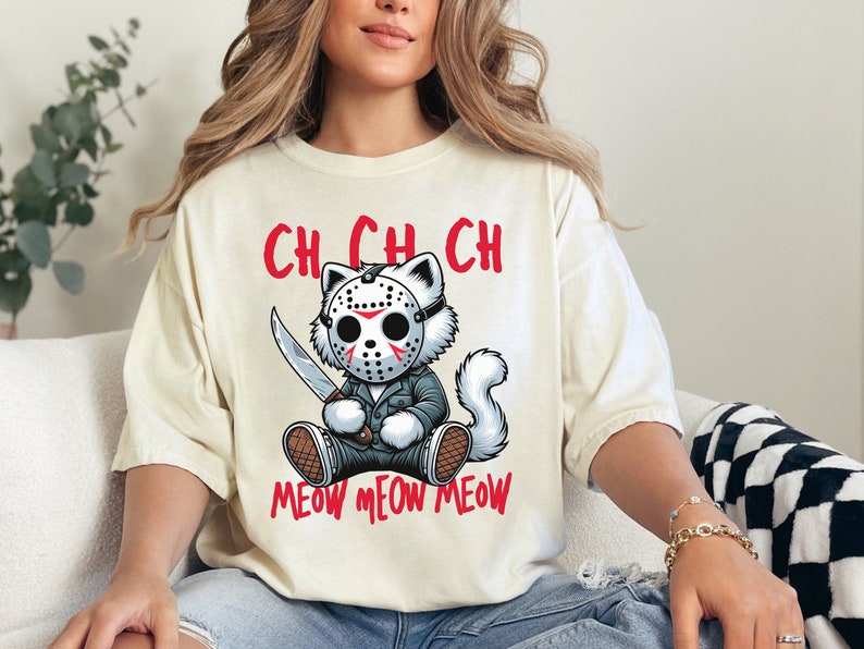 May include: A cream-colored t-shirt with a cartoon cat wearing a hockey mask and holding a knife. The text "Ch Ch Ch Meow Meow Meow" is printed on the shirt.