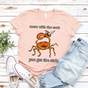 K&ouml;nnte beinhalten: Ein pinkfarbenes T-Shirt mit einem Cartoon-Krebs, der ein Messer h&auml;lt. Der Text auf dem Shirt lautet "mess with the crab you get the stab".