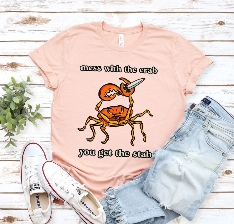 K&ouml;nnte beinhalten: Ein pinkfarbenes T-Shirt mit einem Cartoon-Krebs, der ein Messer h&auml;lt. Der Text auf dem Shirt lautet "mess with the crab you get the stab".