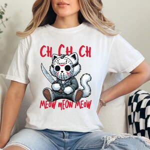 May include: White t-shirt with a graphic of a cat wearing a hockey mask and holding a knife. The text "Ch Ch Ch Meow Meow Meow" is printed below the cat.
