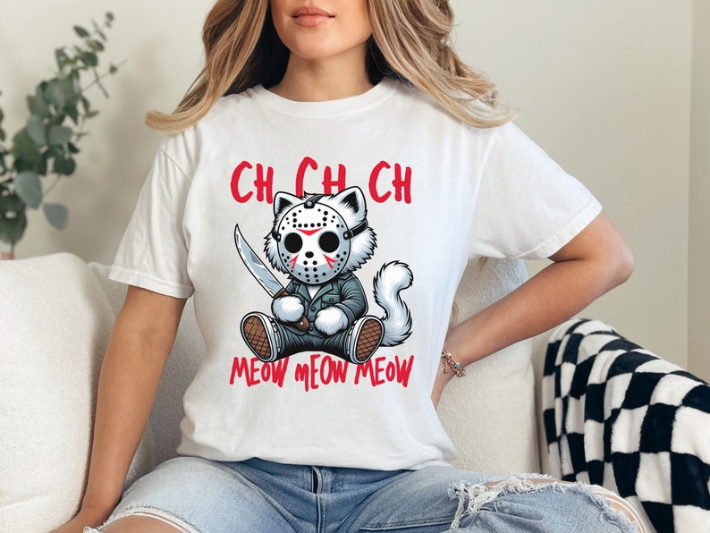 May include: White t-shirt with a graphic of a cat wearing a hockey mask and holding a knife. The text "Ch Ch Ch Meow Meow Meow" is printed below the cat.