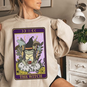 May include: A beige sweatshirt with a purple and gold tarot card design featuring a frog wearing a witch's hat and holding a cup. The card reads "The Witch" and has a crescent moon symbol at the top.