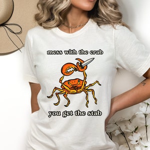 K&ouml;nnte beinhalten: Wei&szlig;es T-Shirt mit einem Grafikdruck eines roten Krebses, der ein Messer h&auml;lt. Der Text "mess with the crab you get the stab" ist auf dem T-Shirt aufgedruckt.