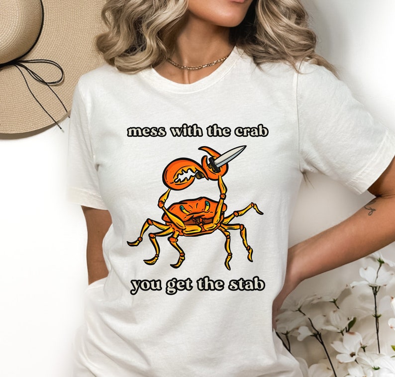 K&ouml;nnte beinhalten: Wei&szlig;es T-Shirt mit einem Grafikdruck eines roten Krebses, der ein Messer h&auml;lt. Der Text "mess with the crab you get the stab" ist auf dem T-Shirt aufgedruckt.