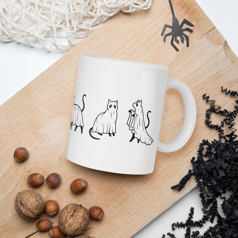 May include: White ceramic mug with a black line drawing of four cats dressed as ghosts. The cats are in various poses, including one holding a ghost sheet.