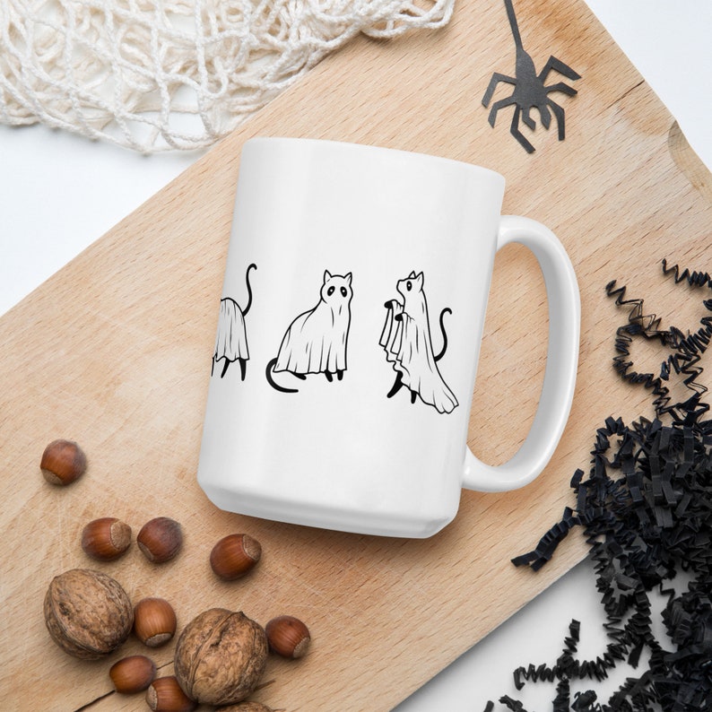 May include: White ceramic coffee mug with a black line drawing of three cats dressed as ghosts. The cats are in various poses, including one holding a ghost sheet.