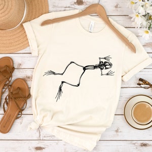 May include: A white t-shirt with a black silhouette of a frog skeleton printed on the front.