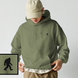 Peut inclure: Un sweat-shirt &agrave; capuche vert avec une petite silhouette noire de Bigfoot marchant sur la poitrine gauche. Le sweat-shirt a une poche kangourou et une capuche &agrave; cordon.