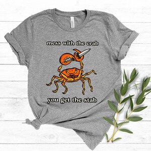 K&ouml;nnte beinhalten: Graues T-Shirt mit einem Cartoon-Krebs, der ein Messer h&auml;lt. Der Text "mess with the crab you get the stab" ist auf das T-Shirt gedruckt.
