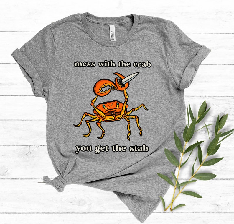 K&ouml;nnte beinhalten: Graues T-Shirt mit einem Cartoon-Krebs, der ein Messer h&auml;lt. Der Text "mess with the crab you get the stab" ist auf das T-Shirt gedruckt.