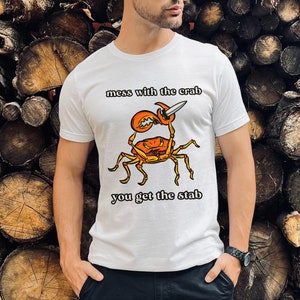 K&ouml;nnte beinhalten: Wei&szlig;es T-Shirt mit einem Grafikdruck eines Krabben, der ein Messer h&auml;lt. Der Text "mess with the crab you get the stab" ist auf dem T-Shirt aufgedruckt.