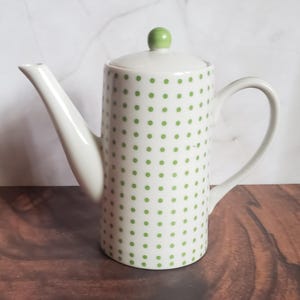 May include: White ceramic teapot with a cylindrical body, a curved handle, and a long spout. The teapot is decorated with evenly spaced green polka dots. The lid has a small green knob. The teapot is on a wooden surface.