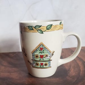 May include: Cream-colored ceramic mug with a birdhouse design. The birdhouse is light blue with red heart accents and two small birds. The top of the mug has a yellow band with green leaves. The mug has a curved handle.