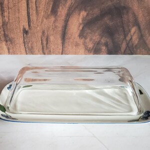 May include: Rectangular glass butter dish with a clear, curved lid. The base is white ceramic with a blue border and floral design. The background is a wooden surface.