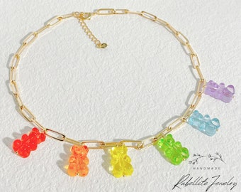 May include: A gold-colored chain necklace featuring colorful gummy bear charms in red, orange, yellow, green, blue, and purple. The necklace has a lobster clasp closure and is displayed on a white surface.