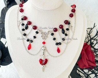 May include: A gothic-style necklace with a silver chain, red and black beads, and a pendant featuring a sword piercing a heart. The necklace has a layered design with dangling teardrop-shaped beads and star charms.