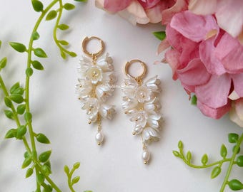May include: A pair of white floral earrings with gold-colored hoops and accents. The earrings feature clusters of small, white flowers and dangling pearls. The background includes pink flowers and green foliage.