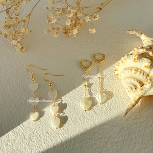 May include: Two pairs of gold-toned earrings. One pair features three white heart-shaped charms on a hook wire. The second pair consists of a small gold hoop with a single white heart-shaped charm suspended from a delicate gold chain.