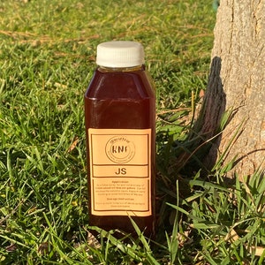 May include: A brown liquid in a clear plastic bottle with a white cap. The label reads "JS" and includes application and storage instructions. The bottle is set on a bed of green grass.