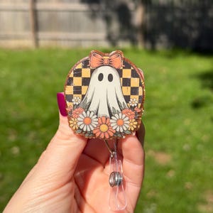 May include: A circular badge reel featuring a white ghost illustration with a peach-colored bow, set against a black and yellow checkered background. The design is surrounded by a border of orange, white, and yellow flowers with glitter accents.
