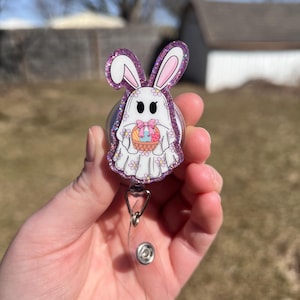 Puede incluir: Un carrete de insignia de purpurina morada con un fantasma blanco con orejas de conejo y una cesta de huevos de Pascua. El carrete es transparente y tiene un clip plateado.
