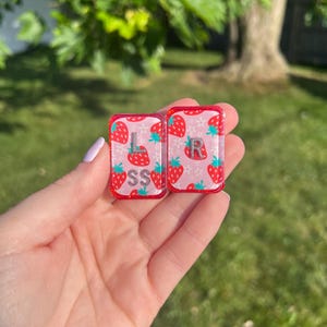 Könnte beinhalten: Zwei rechteckige Acryl-Anhänger mit rosa Hintergrund und einem wiederkehrenden Muster aus roten Erdbeeren und kleinen weißen Blüten. Der linke Anhänger zeigt die silbernen Buchstaben L und SS, der rechte den Buchstaben R.