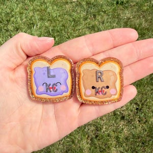 May include: Two toast-shaped pins with glittery borders. The left pin features purple spread with the letters L and KC, while the right pin features tan spread with the letters R and KC. Both have smiling faces with pink cheeks.