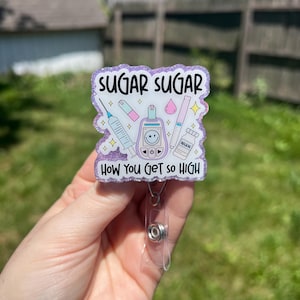 以下が含まれることがあります： SUGAR SUGAR HOW YOU GET SO HIGHという文字が書かれた白いグラフィックと、紫色のグリッターが施されたバッジリール。デザインには、注射器、スマイリーフェイス付きの血糖値測定器、ピンクのしずく、ペン、インスリン瓶のイラストが含まれています。