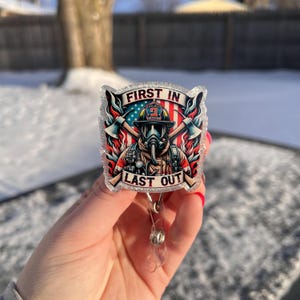 Könnte beinhalten: Ein Ausweis-Rollhalter mit einem Feuerwehr-Motiv und dem Text „FIRST IN LAST OUT“. Das Design zeigt einen Feuerwehrmann mit Gasmaske, Äxten und der amerikanischen Flagge. Das Rollhalter ist transparent mit Glitzer.