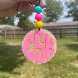 May include: A pink acrylic car charm with a glittery background and the letters GSR. The charm is round and has a black string with three colorful beads: teal, pink, and yellow. The background is a blurred outdoor scene.