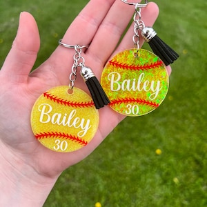 May include: Two softball-themed keychains. Each keychain is round, yellow, and glittery, with red stitching details. The name "Bailey" and the number "30" are printed on each keychain. Each keychain has a black tassel and a silver keyring.