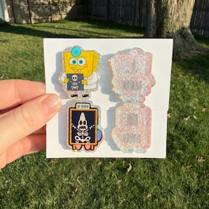 May include: Two acrylic pins featuring cartoon characters. One pin shows a yellow sponge character in a white lab coat holding an X-ray. The other pin shows a pink starfish character skeleton inside a frame labelled X-RAY. Both have glittery finishes.