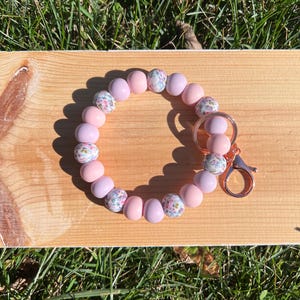 Pulsera con flores en tonos pastel, pulsera con cuentas de silicona con purpurina, llavero en tono rosa pastel, pulsera con cuentas en tono morado pastel, pulsera con llavero en tono oro rosa