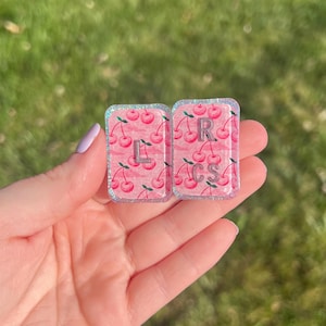 Puede incluir: Dos dijes rectangulares de resina con fondo rosa, un patrón de cerezas repetido y un borde brillante. El dije izquierdo muestra la letra L en plata, mientras que el dije derecho muestra las letras R y CS en color plata.