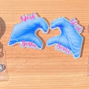 May include: Two retractable badge holders and two blue glove-shaped designs forming a heart. The designs have the words "Work Bestie" in pink glitter. The badge holders have clear plastic straps and silver accents.