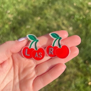 Peut inclure: Deux broches ou breloques en acrylique en forme de cerise. La cerise de gauche porte les lettres L et AS en argent, tandis que la cerise de droite porte la lettre R. Les deux cerises sont rouge vif avec des tiges vertes et une texture pailletée.