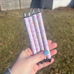 Peut inclure: Quatre stylos avec un motif blanc et lavande représentant des lapins, des carottes et des fleurs. Les stylos ont des pointes argentées et de l'encre noire. Les stylos sont tenus dans une main avec du vernis à ongles bleu. L'arrière-plan est flou.