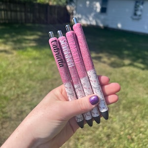 May include: Four pink glitter pens with names: Gabriella, Savannah, and Elizabeth. The pens have a white base with a ribbon and heart pattern. The pens are held in a hand.