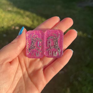 May include: Two rectangular pink glitter resin charms featuring black skeleton hand illustrations. The left charm displays the letters L and RM, while the right charm displays the letters R and RM in a silver metallic font against the pink glitter.