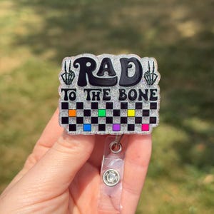 May include: A silver glitter badge reel featuring the black text RAD TO THE BONE with skeleton hand peace signs on the sides. Below the text is a black and white checkered pattern with small colorful squares in orange, green, yellow, blue, purple, and pink.