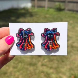 May include: A pair of ghost-shaped earrings featuring a black base with neon pink, blue, and orange outlines, topped with a bow. The left earring has the letters L and RT printed on it, while the right earring features the letter R in silver.