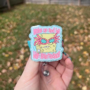 May include: A light blue glitter badge reel featuring a cartoon character with pink glasses and the text "Wake up and be High Maintenance" in a glittery font. The reel has a clear plastic retractable cord and a silver clip.