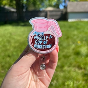 May include: A pink and glittery badge reel in the shape of a coffee pot with the text "Pour Myself a Cup of Ambition" in blue and white. The badge reel has a clear plastic retractable cord and a silver-colored clip.