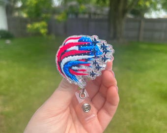 May include: A badge reel with a patriotic design. The badge reel features a stylized American flag with red, white, and blue colors and stars. The reel has a clear plastic retractable cord and a clip for attaching to clothing.