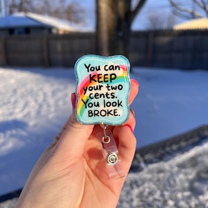 May include: A badge reel with a light blue background and a rainbow design. The text on the badge reel reads "You can KEEP your two cents. You look BROKE."