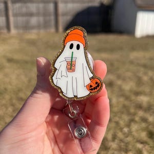 May include: A white ghost with a pumpkin and a cup of coffee, wearing an orange hat, is attached to a clear plastic badge holder with a silver clip.
