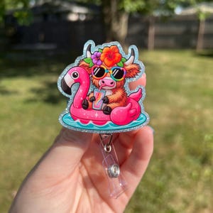May include: A glittery badge reel featuring a cartoon Highland cow wearing sunglasses and a flower crown, floating on a pink flamingo pool float. The cow is holding a drink with a rainbow straw. The badge reel has a clear plastic clip.