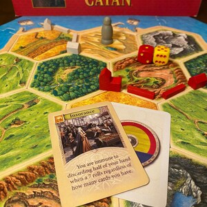 May include: A Settlers of Catan board game set featuring a hexagonal terrain board, red and white wooden player pieces, a grey pawn, two dice, and an Insurance game card with text stating you are immune to discarding half of your hand.
