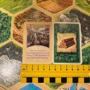 May include: Two rectangular game cards placed above a yellow 14 cm ruler. The left card is titled Insurance and features an illustration of people in a market, with text describing immunity to discarding cards. The right card shows a stack of logs.