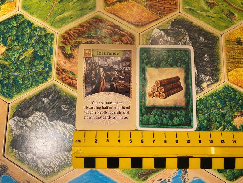 May include: Two rectangular game cards placed above a yellow 14 cm ruler. The left card is titled Insurance and features an illustration of people in a market, with text describing immunity to discarding cards. The right card shows a stack of logs.
